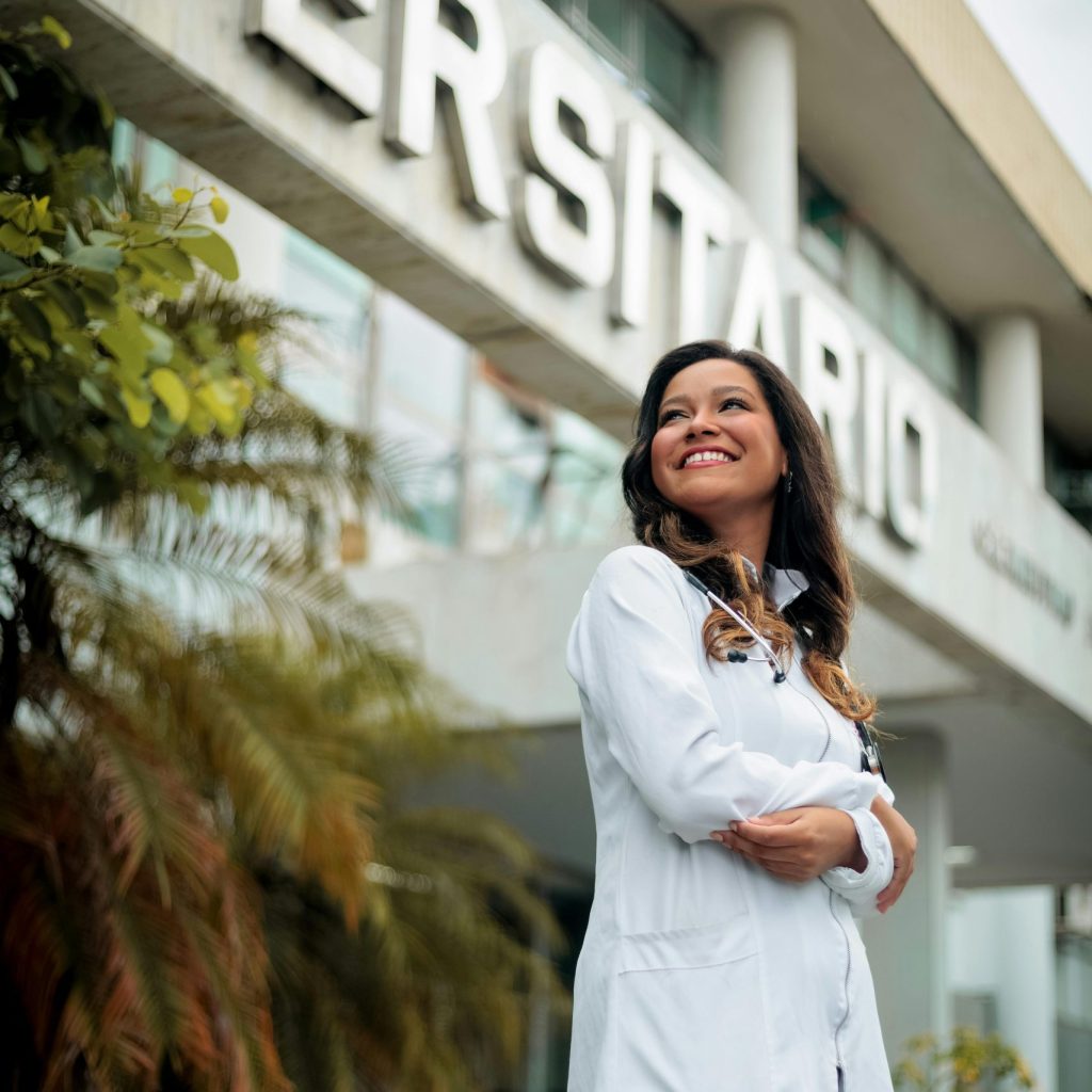 Smiling female doctor in white coat standing outdoors near university building, expressing confidence and optimism.